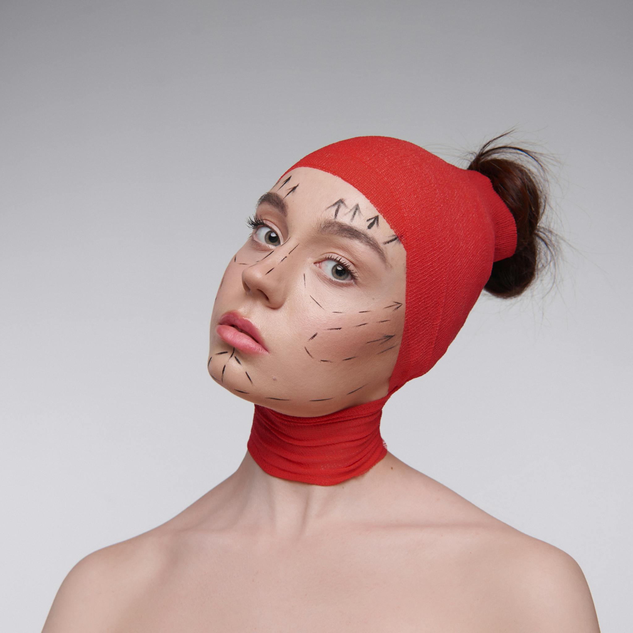 Creative portrait of a woman with surgical markings wearing a red head and neck wrap, illustrating beauty concepts.
